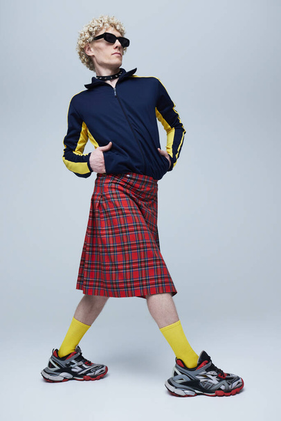 Retrato de cuerpo entero del joven hombre de pelo rizado posando en escocés escocés y ropa deportiva sobre fondo gris estudio. Concepto de fusión de moda, estilo, queer, singularidad, auto-expresión. Anuncio - Foto, Imagen