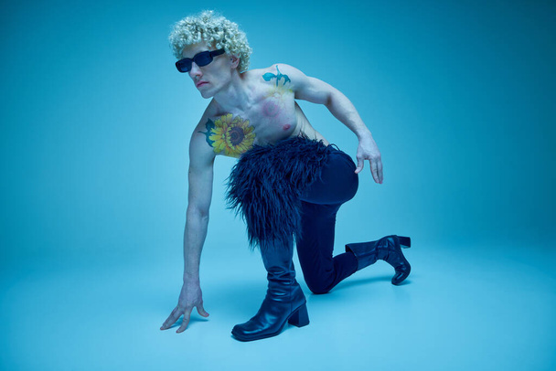 Πορτρέτο του ανδρικού μοντέλου σε εντυπωσιακή μόδα-προς τα εμπρός στάση χορού, ανάμειξη γοητεία και εναλλακτικό στυλ σε μπλε φόντο. Έννοια της σύντηξης μόδας, φωτογραφία τέχνης, στυλ, queer, αυτο-έκφραση. - Φωτογραφία, εικόνα