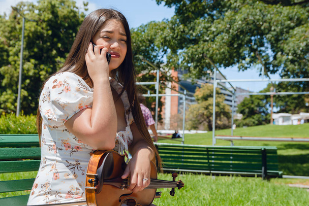 νεαρή Λατίνα βιολιστής με μακριά μαλλιά και φόρεμα, κάθεται με το βιολί της στα πόδια της, μιλώντας στο τηλέφωνο στο πάρκο το μεσημέρι. - Φωτογραφία, εικόνα