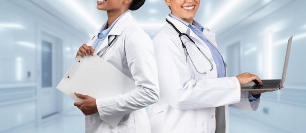 Két örömteli egészségügyi szakember, az egyik írótáblával, a másik laptoppal, háttal áll a kórházi folyosón, bemutatva a csapatmunkát az egészségügyben a klinikán, panoráma - Fotó, kép