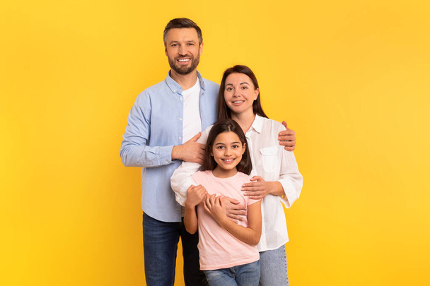 Liefdevol Family Concept. Millennial ouders en hun dochter poseren in tedere knuffel, gekleed in casual kleding, stralend geluk in familieportret over gele achtergrond - Foto, afbeelding