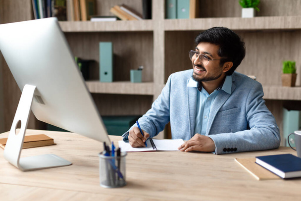 Empresário indiano sorrindo trabalhando na mesa com computador e tomando notas, planejando seu trabalho diurno no interior do escritório moderno. Empreendedor do Oriente Médio bem sucedido escrevendo relatório de negócios perto do PC - Foto, Imagem