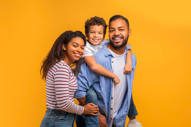 Οικογενειακό πορτρέτο. Happy Young Black Parents and cute Preteen Son Embracing Together, Χαρούμενη Αφρο-Αμερικανίδα μητέρα, πατέρας και αρσενικό παιδί ποζάρουν ενάντια φωτεινό κίτρινο φόντο στούντιο, αντίγραφο χώρου - Φωτογραφία, εικόνα
