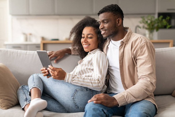 Happy black millennial couple browsing online together on digital tablet, boyfriend and girlfriend relaxing with gadget on couch, watching film online, enjoying modern lifestyle at home - Photo, Image