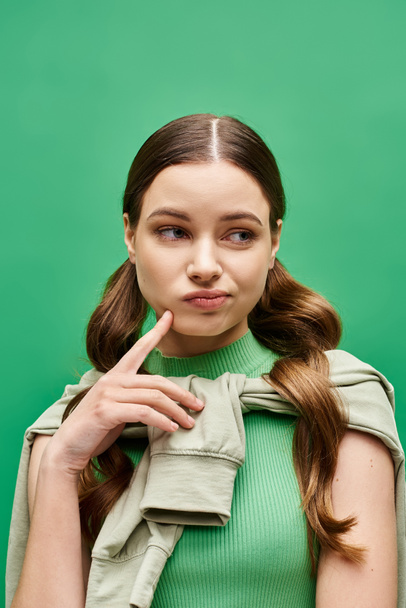 20代のスタイリッシュな若い女性がスタジオの設定で頬を指差し,印象的な緑のセーターを着ています. - 写真・画像