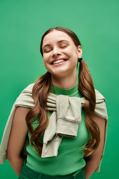 Μια χαρούμενη νεαρή γυναίκα, γύρω στα 20, με μακριά μαλλιά, ποζάρει με χάρη σε ένα πράσινο πουλόβερ σε ένα στούντιο.. - Φωτογραφία, εικόνα