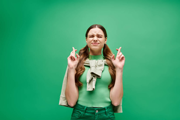 Eine junge Frau in ihren Zwanzigern drückt in einem Studio vor grünem Hintergrund die Daumen vor ihrem Gesicht. - Foto, Bild