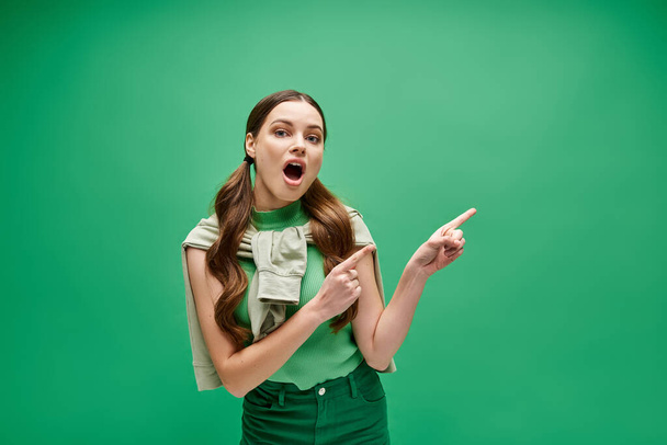 Młoda kobieta po dwudziestce, ubrana w zieloną koszulę, wskazuje na coś poza ekranem z dziwnym wyrazem twarzy.. - Zdjęcie, obraz