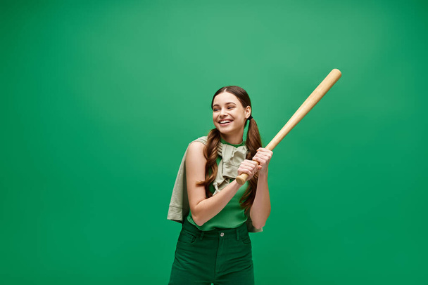 Een jonge vrouw van in de twintig houdt een honkbalknuppel vol vertrouwen tegen een levendige groene achtergrond, straalt kracht en vastberadenheid uit.. - Foto, afbeelding