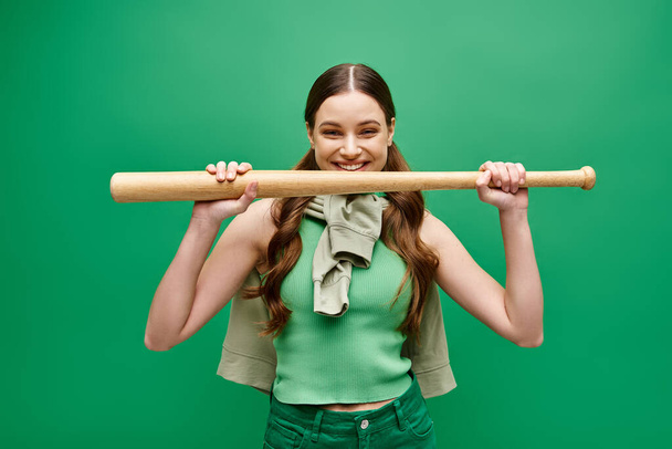Una joven y hermosa mujer de unos 20 años sostiene un bate de béisbol sobre su cabeza en una pose dinámica contra un fondo de estudio verde. - Foto, Imagen