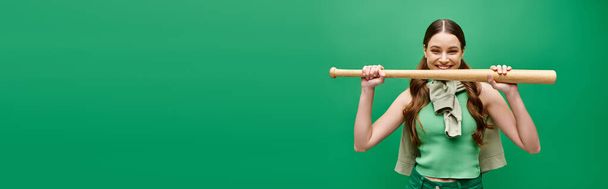 Een jonge, mooie vrouw van in de twintig houdt een honkbalknuppel voor haar gezicht in een studio op groen. - Foto, afbeelding