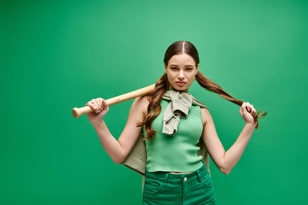 Молодая женщина лет 20 уверенно держит бейсбольную биту над плечом в студии на зеленом фоне.. - Фото, изображение