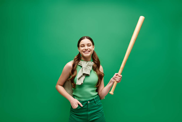 Μια νεαρή γυναίκα γύρω στα 20 κρατά με αυτοπεποίθηση ένα ρόπαλο του μπέιζμπολ σε ένα εντυπωσιακό πράσινο φόντο. - Φωτογραφία, εικόνα