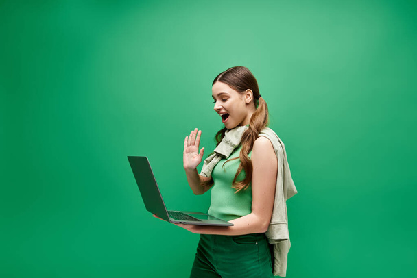 Una joven de unos 20 años, vestida con un vestido verde vibrante, sostiene un portátil en un entorno de estudio moderno. - Foto, imagen