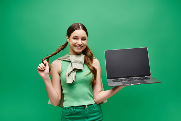 Eine junge Frau in ihren Zwanzigern hält selbstbewusst einen Laptop in der Studiokulisse mit lebendigem grünen Bildschirmhintergrund. - Foto, Bild
