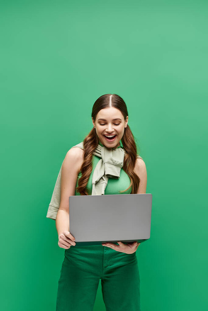 Egy húszas éveiben járó fiatal nő áll egy zöld fal előtt, magabiztosan tartva egy laptopot.. - Fotó, kép