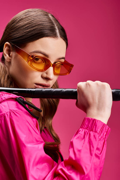 ピンクのジャケットを着た20代のスタイリッシュな若い女性は,鮮やかなピンクの背景に対して自信を持って野球バットを保持しています. - 写真・画像