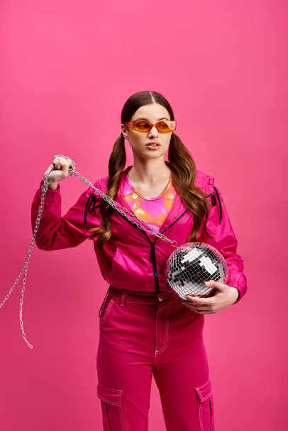 Uma mulher vibrante de 20 anos, vestindo uma roupa rosa elegante, segura uma bola de discoteca, exalando energia contra um pano de fundo rosa. - Foto, Imagem