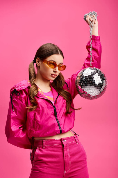 Стильная молодая женщина в возрасте 20 лет, одетая в ярко-розовый костюм, держащая мерцающий диско-шар в студии с розовым фоном. - Фото, изображение