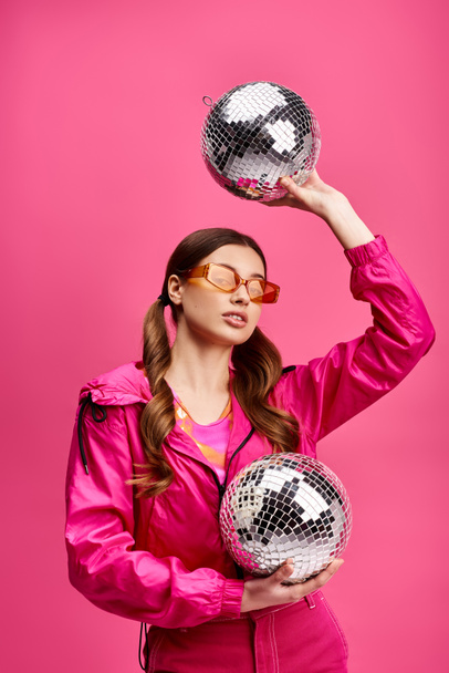 Mladá žena ve věku 20 let, stylový v růžové bundě, drží dvě disco koule na pulzujícím růžovém pozadí. - Fotografie, Obrázek