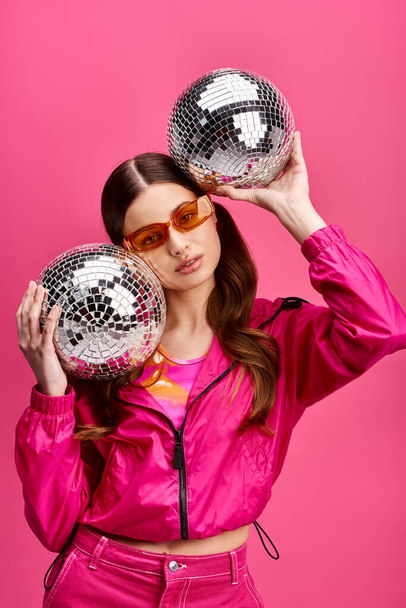 A stylish woman in her 20s, clad in a pink jacket, holds two disco balls in a studio with a vibrant pink background. - Photo, Image