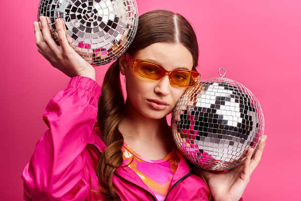 ピンクのジャケットを着ている20代の若くてスタイリッシュな少女は,ピンクの背景を持つスタジオで2つのディスコボールを保持しています.. - 写真・画像