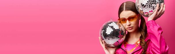Uma jovem mulher elegante na casa dos 20 anos, vestindo uma camisa rosa, alegremente segurando duas bolas de discoteca em um estúdio com um fundo rosa. - Foto, Imagem