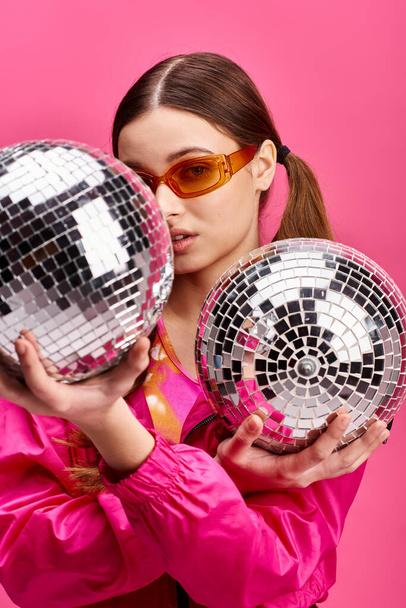 Une jeune femme élégante dans la vingtaine tient deux boules de disco dans un studio avec un fond rose, respirant une ambiance glamour. - Photo, image