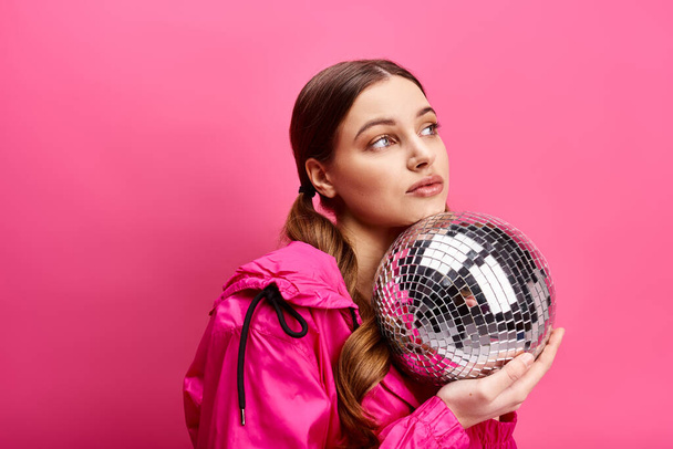 Een stijlvolle jonge vrouw in de 20 met een roze jasje, met een sprankelende discobal in een studio met een roze achtergrond. - Foto, afbeelding