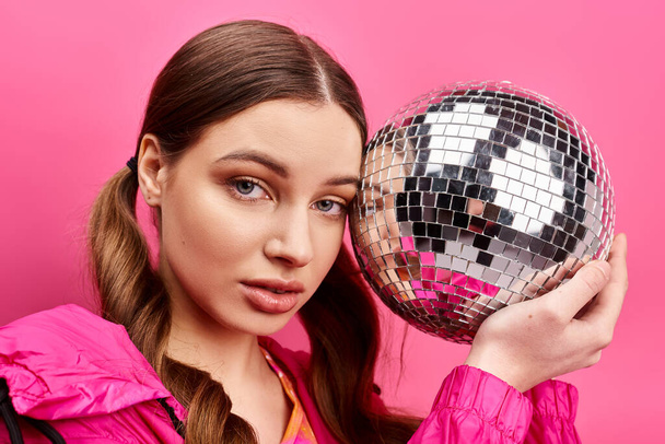 Een stijlvolle jonge vrouw van in de twintig houdt een discobal voor haar gezicht en creëert een betoverende reflectie. Roze achtergrond in studio. - Foto, afbeelding