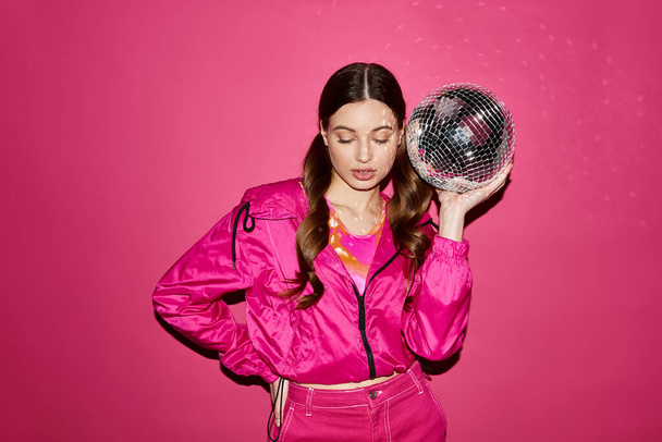 Μια νεαρή γυναίκα γύρω στα 20, κομψή με ροζ μπουφάν, κρατά μια ντίσκο μπάλα σε ένα στούντιο με ένα ζωντανό ροζ φόντο. - Φωτογραφία, εικόνα