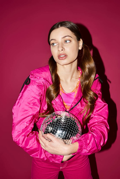 Uma jovem elegante de 20 anos usando uma jaqueta rosa segura uma bola de discoteca em um estúdio com um fundo rosa. - Foto, Imagem