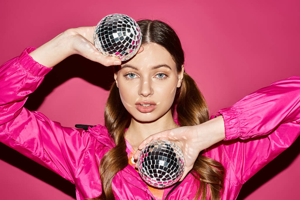 Una mujer joven y elegante de unos 20 años sosteniendo dos bolas de discoteca en un estudio con un fondo rosa. - Foto, imagen