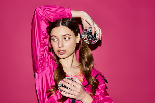 Una elegante joven de unos 20 años con un atuendo rosa sostiene una deslumbrante bola de discoteca en un estudio con un fondo rosa. - Foto, Imagen