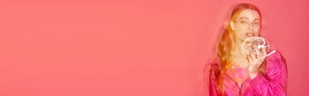 Молодая женщина в розовом платье элегантно держит диско-шар, создавая мечтательную и волшебную атмосферу в студии с розовым фоном. - Фото, изображение
