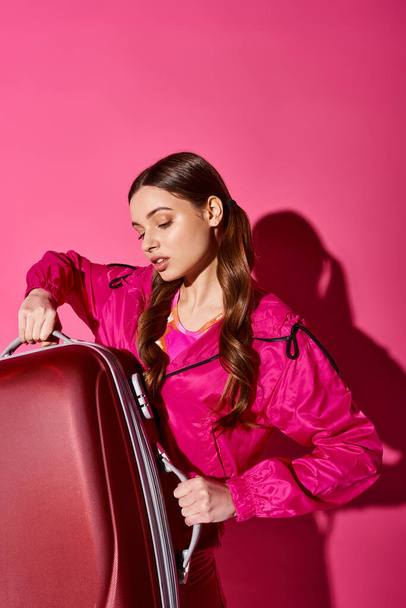 Μια κομψή γυναίκα γύρω στα 20 ντυμένη στα ροζ κρατά μια βαλίτσα σε ένα στούντιο με ροζ φόντο, αποπνέοντας μια αίσθηση περιπέτειας. - Φωτογραφία, εικόνα