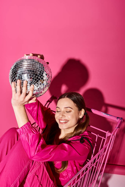 Een jonge vrouw in een stijlvolle roze outfit met een discobal, die glamour en plezier uitstraalt tegen een levendige roze achtergrond. - Foto, afbeelding