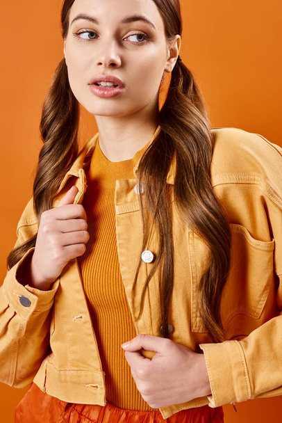 Een jonge, stijlvolle vrouw van in de twintig met lang haar, in een geel jasje, poserend in een studio tegen een oranje achtergrond. - Foto, afbeelding