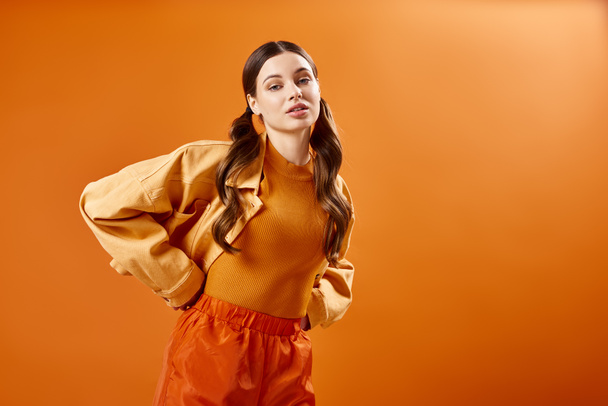 Modna młoda kobieta po dwudziestce z pewnością pozuje w stylowym pomarańczowym stroju na tętniącym życiem tle studia. - Zdjęcie, obraz