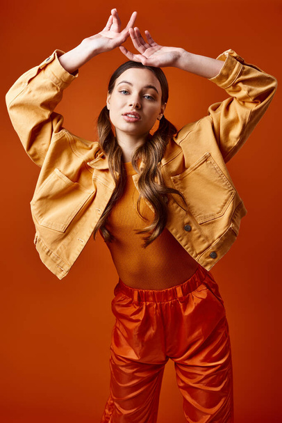スタジオの設定で明るいオレンジ色の背景に黄色のジャケットとオレンジ色のズボンを着用した20代のスタイリッシュな女性. - 写真・画像