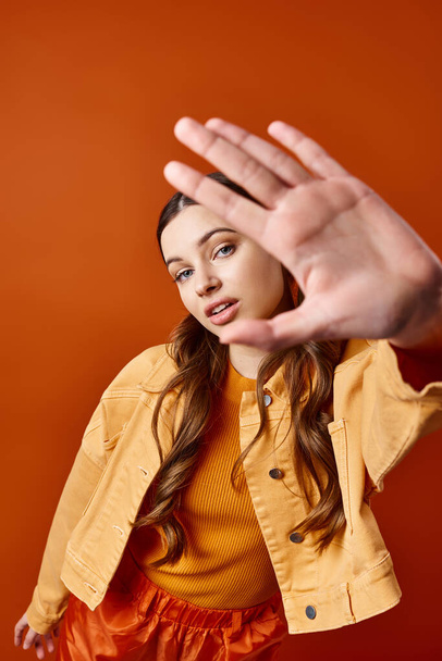 Eine stilvolle Frau in ihren Zwanzigern in einer gelben Jacke macht vor orangefarbenem Hintergrund in einem Atelier eine auffällige Handgeste. - Foto, Bild