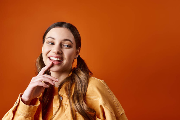 Una mujer joven y elegante de unos 20 años posa con el dedo en los labios contra un fondo de estudio naranja. - Foto, imagen