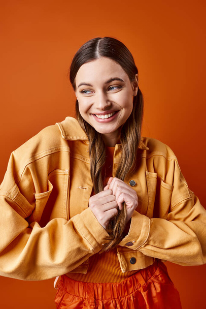 Una mujer joven y elegante de unos 20 años con el pelo largo y una chaqueta amarilla vibrante sobre un fondo de estudio naranja. - Foto, Imagen