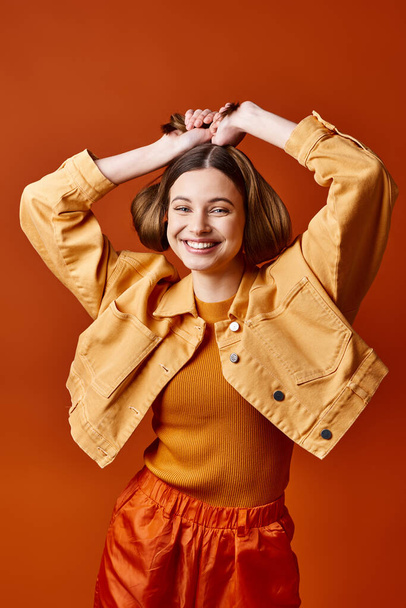 Fiatal, stílusos nő a 20-as éveiben, élénk sárga kabátot és narancssárga nadrágot visel, egy narancssárga hátterű stúdióban pózol. - Fotó, kép