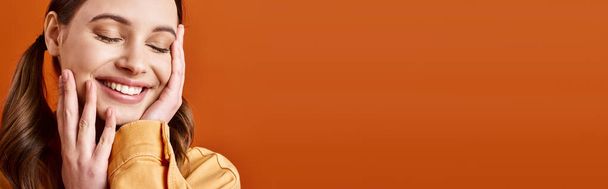 Een jonge, stijlvolle vrouw in de 20 met handen op het gezicht, diep in gedachten, tegen een oranje studio achtergrond. - Foto, afbeelding