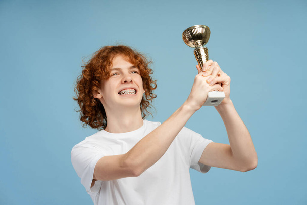Portret van een positieve tiener met een tandbeugel die de beker op een blauwe achtergrond vasthoudt en wegkijkt. Begrip overwinning, competitie  - Foto, afbeelding