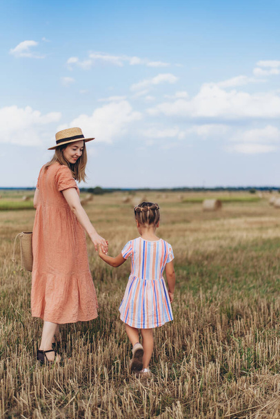 Uma jovem mãe em um vestido longo e um chapéu de palha leva uma pequena filha pela mão. Ambos caminham através de um campo de trigo colhido. Contra o fundo de um fardo de palha - Foto, Imagem