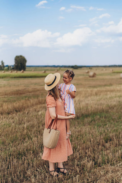 Uma jovem mãe em um vestido longo e um chapéu de palha leva uma pequena filha pela mão. Ambos caminham através de um campo de trigo colhido. Contra o fundo de um fardo de palha - Foto, Imagem