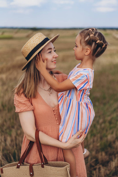 Молодая мать в соломенной шляпе и с плетеной сумкой держит на руках маленькую дочь. Оба стоят в поле собранной пшеницы. На фоне тюка соломы - Фото, изображение