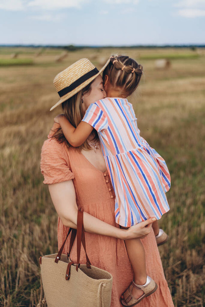 Una giovane madre con un cappello di paglia e con una borsa di vimini tiene una piccola figlia tra le braccia. Entrambi si trovano in un campo di grano raccolto. Sullo sfondo di una balla di paglia - Foto, immagini